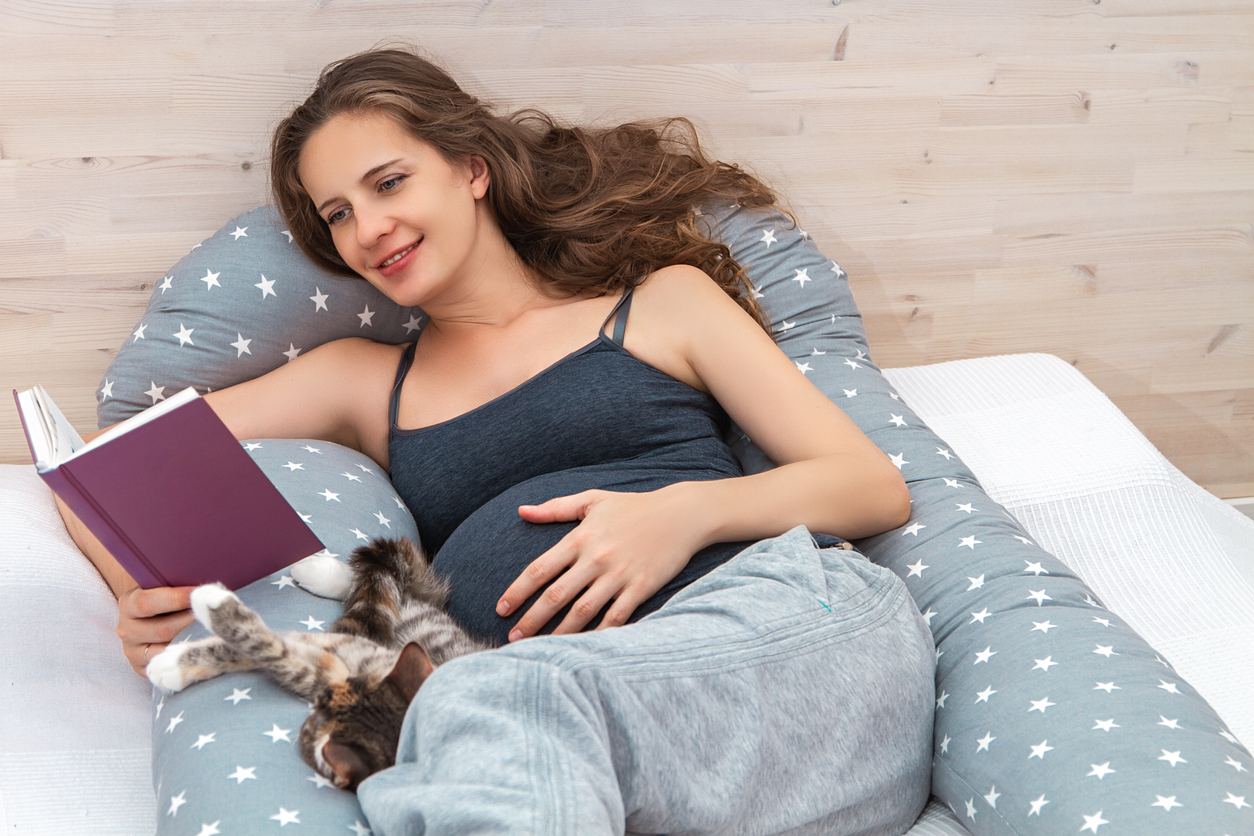 Beautiful pregnant woman lying on bed on body pillow indoor with cat, reading book and touching her tummy.