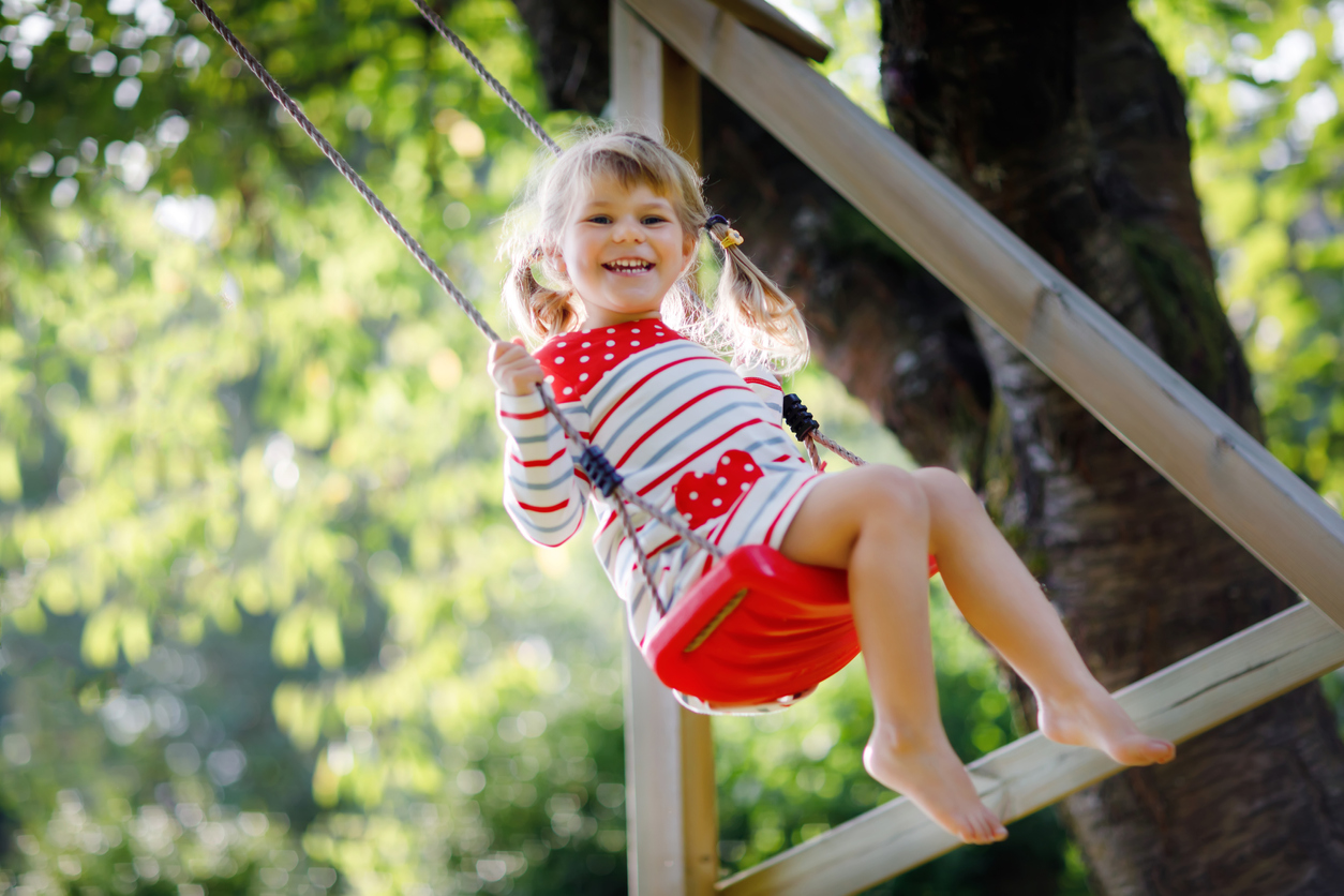 Happy beautiful little toddler girl having fun on swing in domestic garden. Cute healthy child swinging under blooming trees on sunny spring day. Baby laughing and crying