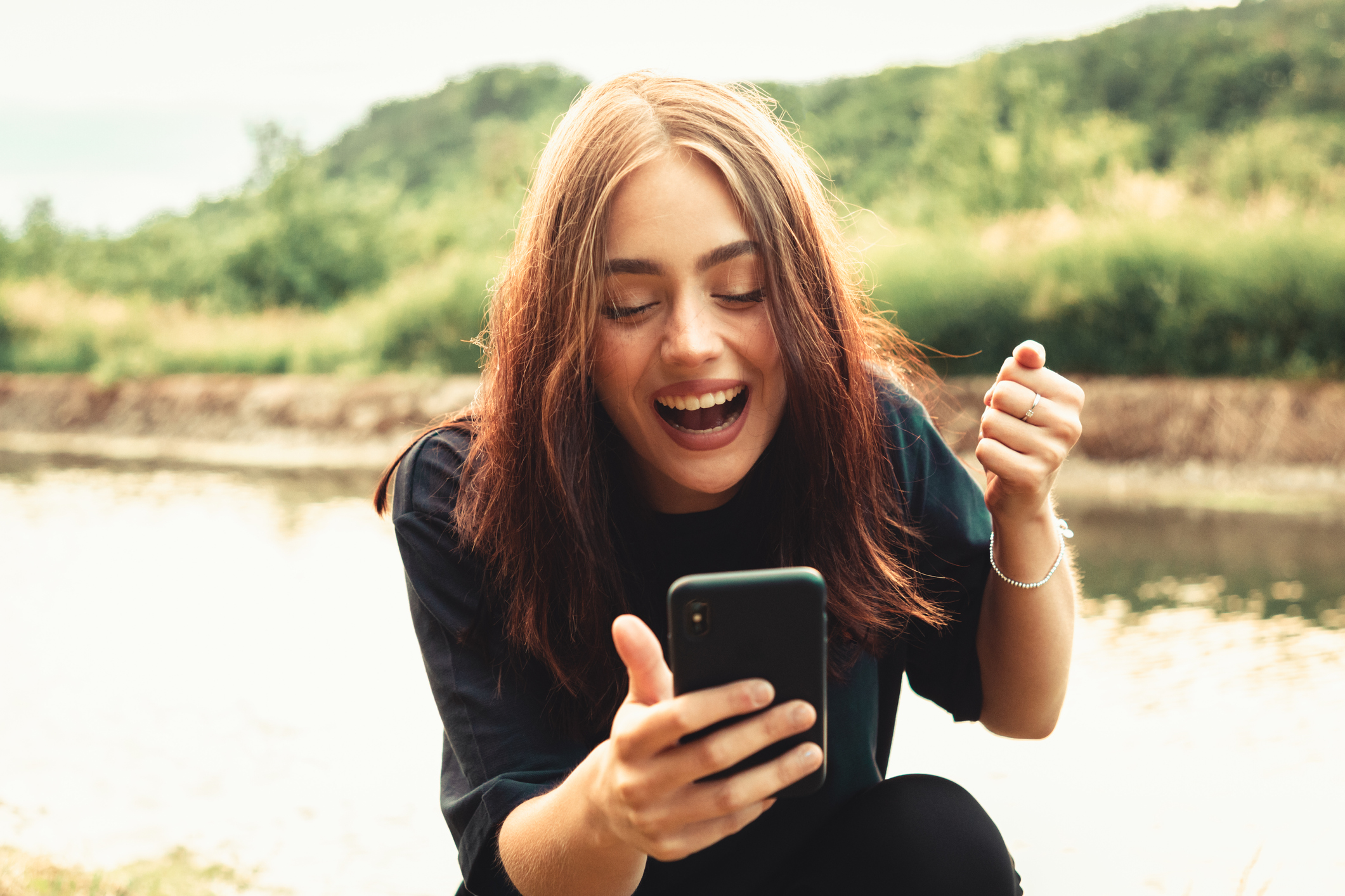 Young smiling woman sitting next to a river typing messages on her smart phone