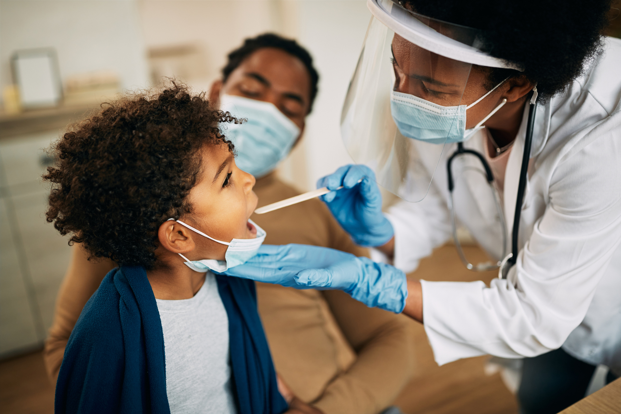 African American doctor with face mask examining boy's throat during a home visit.