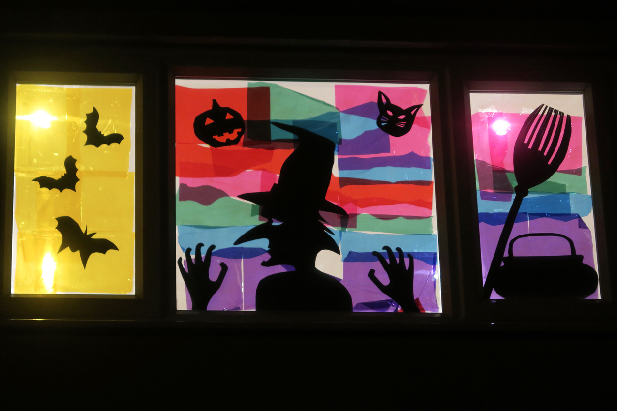 Image of Halloween scene window picture, back lit witch silhouette with bats, cauldron, broom, pumpkin and cat depicted on window pane, tissue paper stained glass effect art