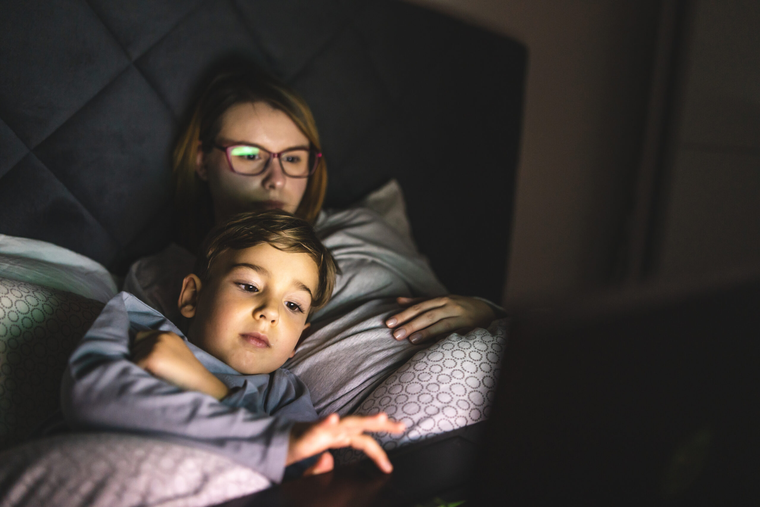 Mother and son watching movie or cartoon using laptop in bed at nighttime - Small boy and his mom using computer while lying in bed at night in dark room - leisure activity family bedtime concept