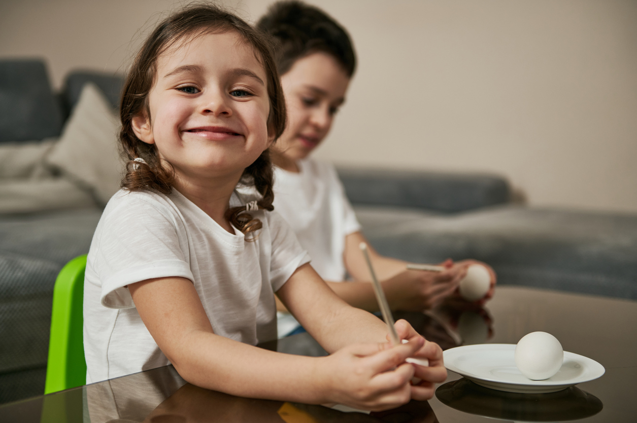 Adorable girl sitting near a school boy, holding a paintbrush and smiling to camera before coloring Easter eggs