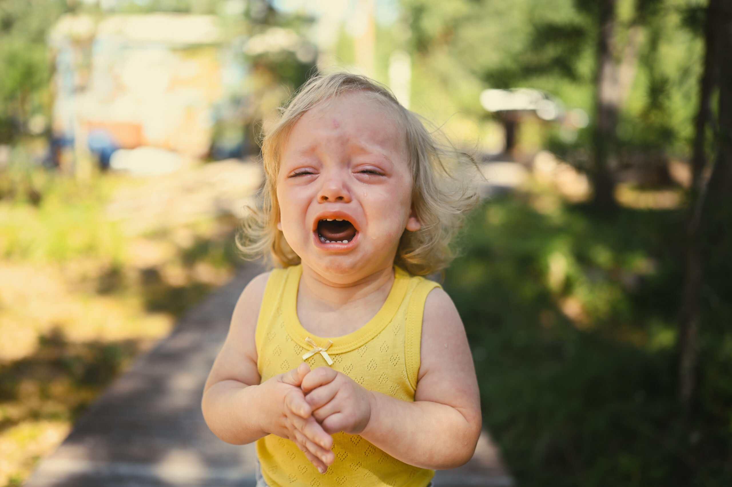 Close up portrait of little funny cute blonde girl child toddler in yellow bodysuit crying outside at summer. Childish tantrum. Healthy childhood concept.