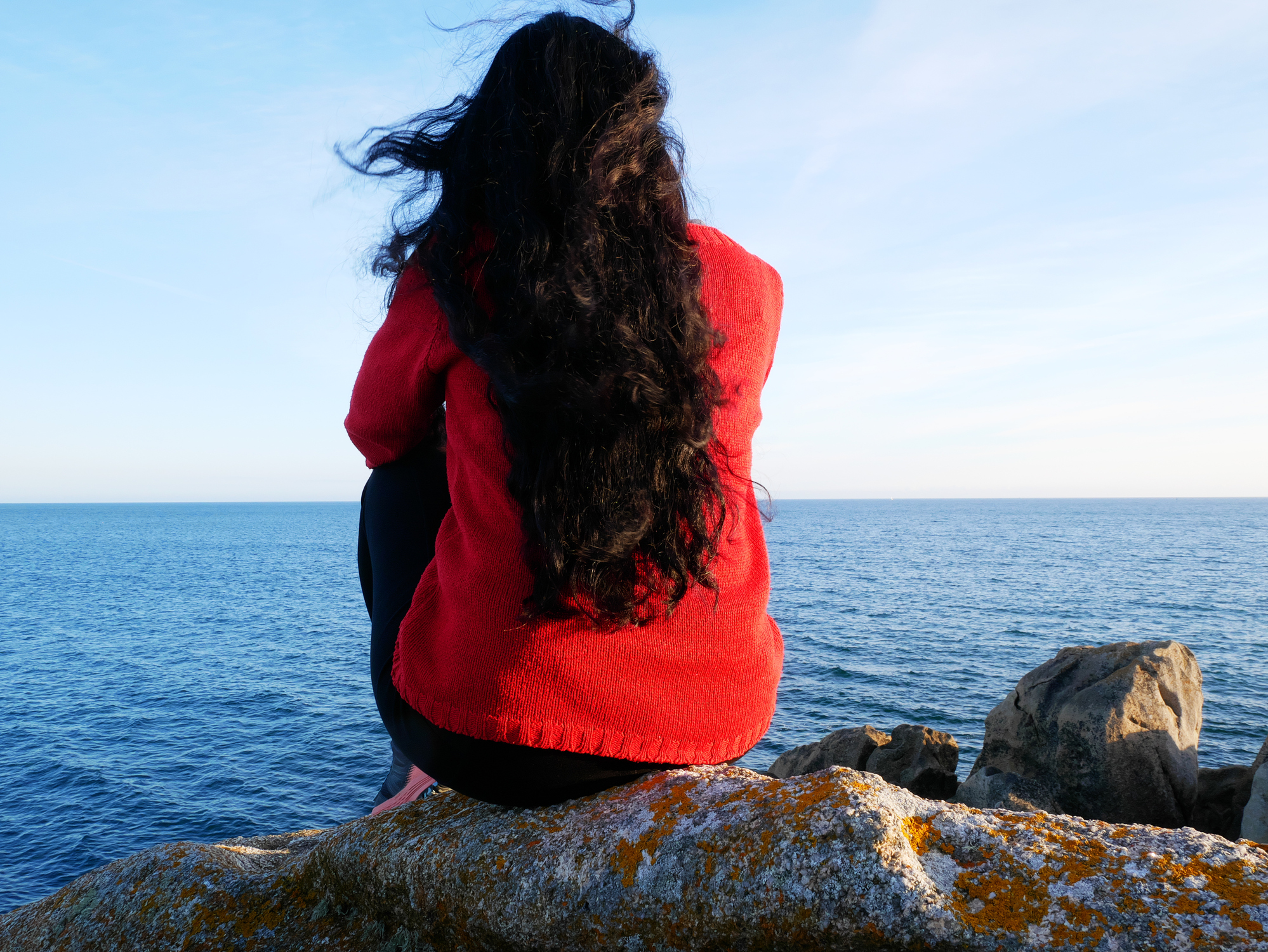 Woman with long black hair, sitting on a rock, at the edge of the ocean