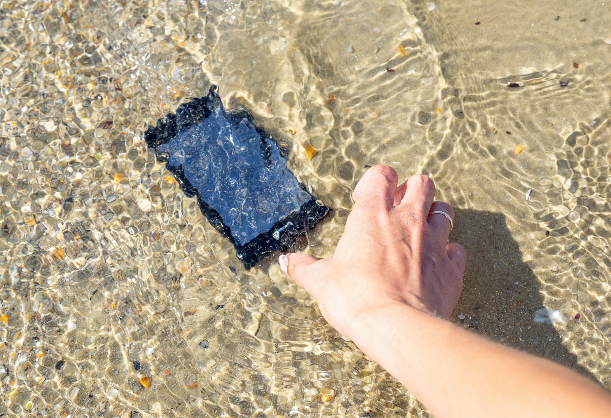 A woman's hand lifts a her smartphone from sea water on the beach.