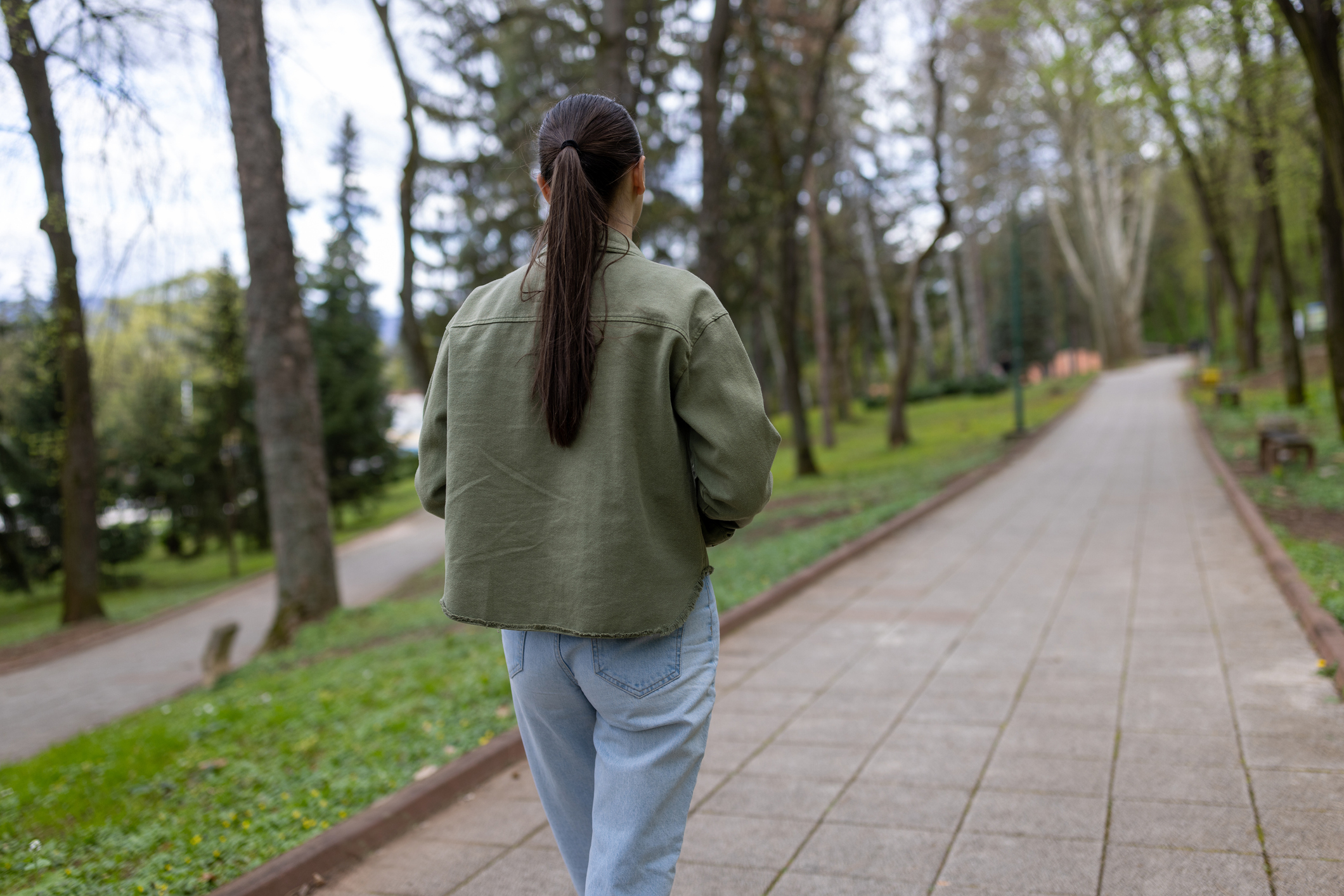 Young casually clothed woman slowly walking through alley of green trees on sidewalk at city park