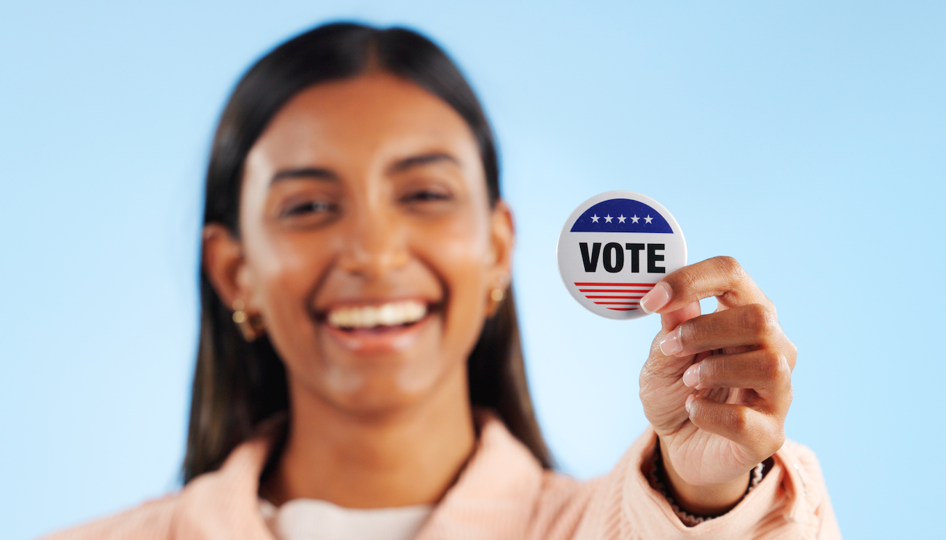 Woman, hand and pin for vote by showing in studio for mock up in politics on blue background. Indian person, smile and excited for government election or campaign with support in community engagement