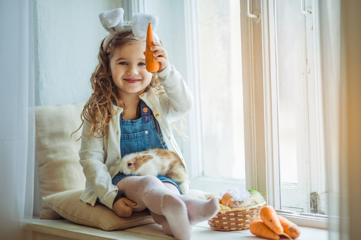 Cute happy little child girl is wearing bunny ears on Easter day holding her friend little rabbit and carrots