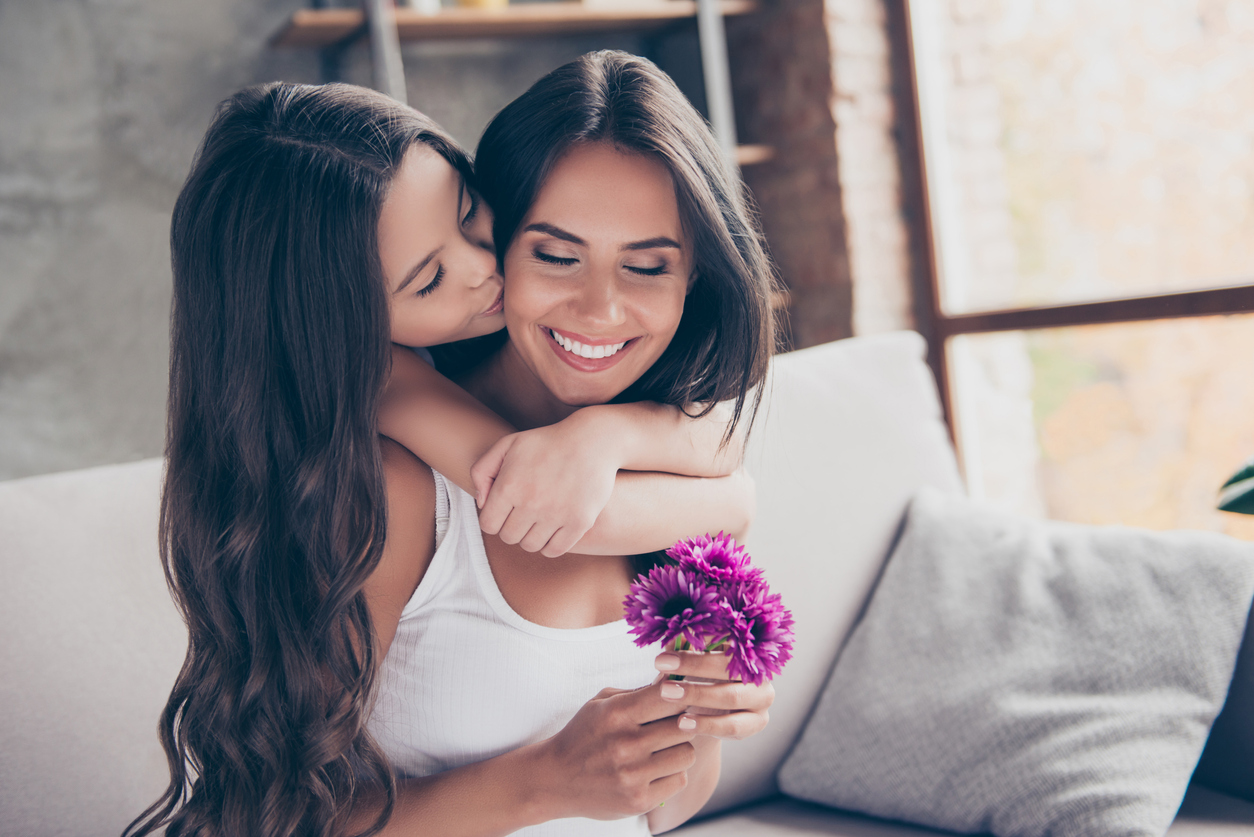 Concept of women's day! Amazed cheerful joyful charming mum is holding nice flowers and is receiving a kiss from her little cute daughter with long curly hair