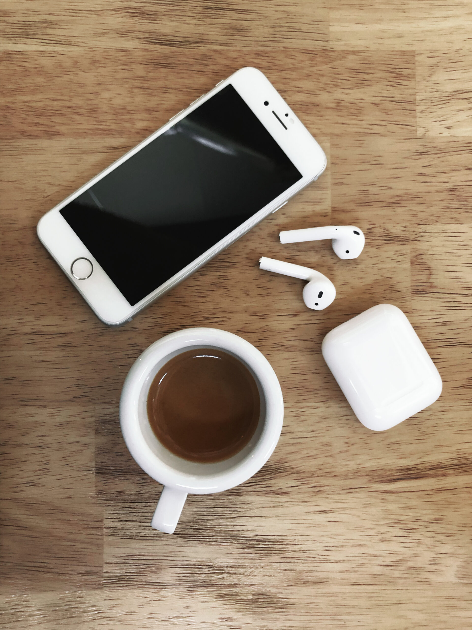 White colored Apple brand Iphone 8 Phone and Airpods 2 on a wooden table with charging box and cup of espresso