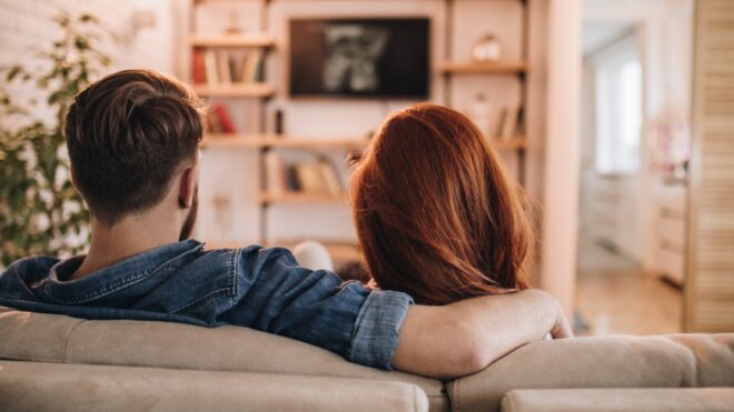 Back view of relaxed couple watching a movie on TV in the living room.