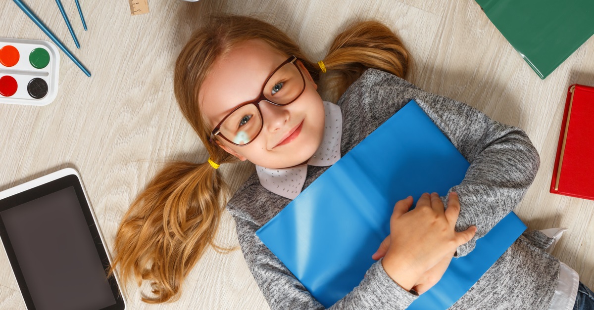 Cute little girl in glasses with a book lying on the floor. A child is surrounded by a book, tablet, paints, brushes, pencils.