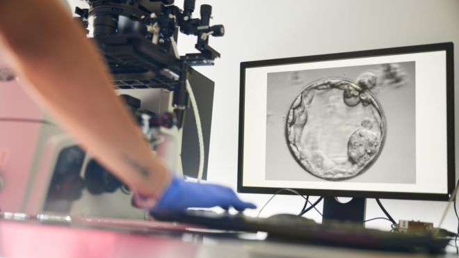 A fertility procedure is shown on the screen while a woman in scrubs follows the process in the lab.