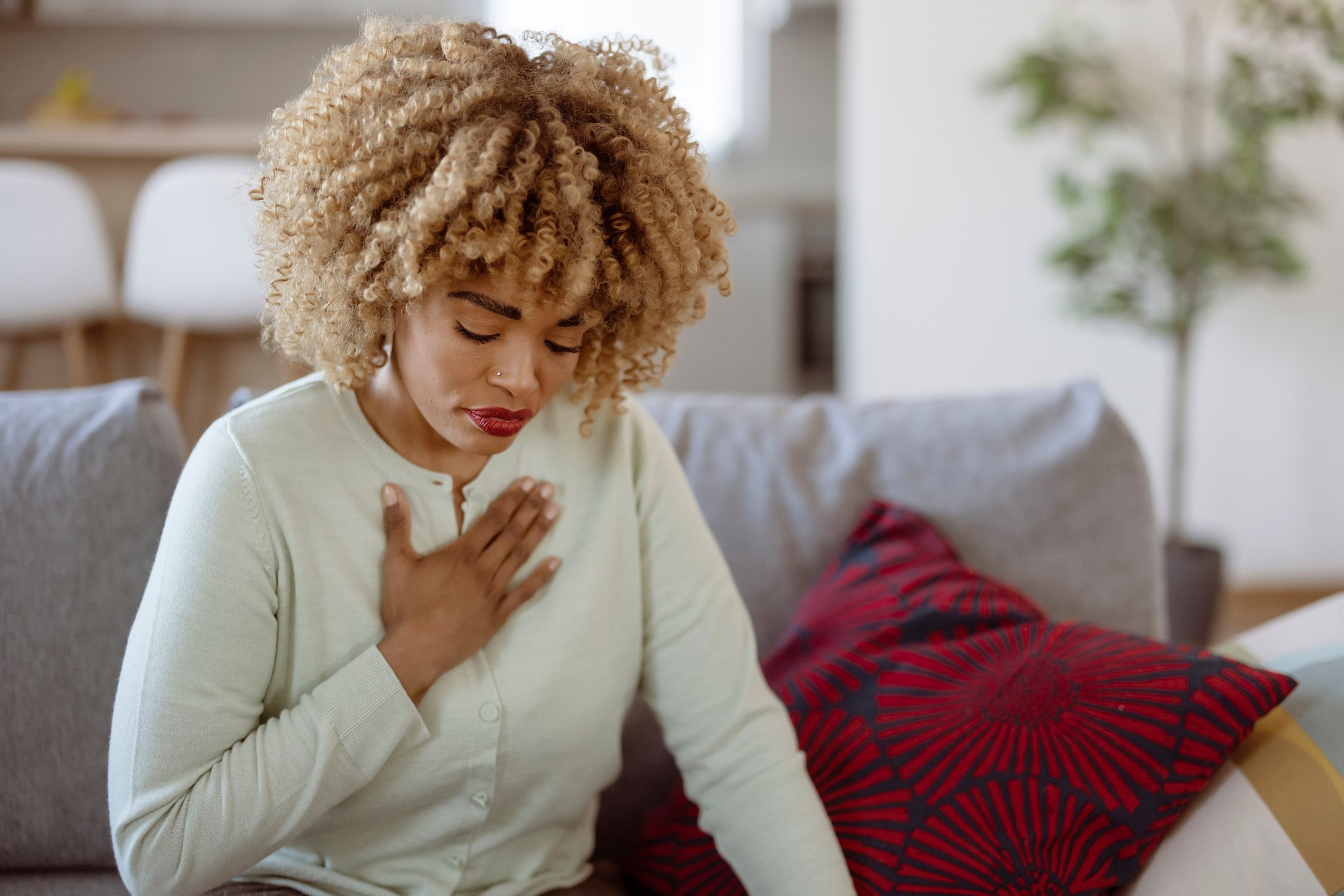 A Concerned Woman Sits on a Gray Couch in a Modern Living Room, Clutching Her Chest With a Distressed Expression