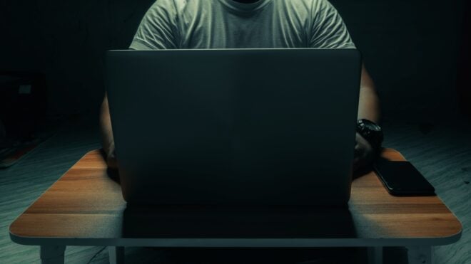 A man in a white shirt sits on a laptop in a dark room. with light