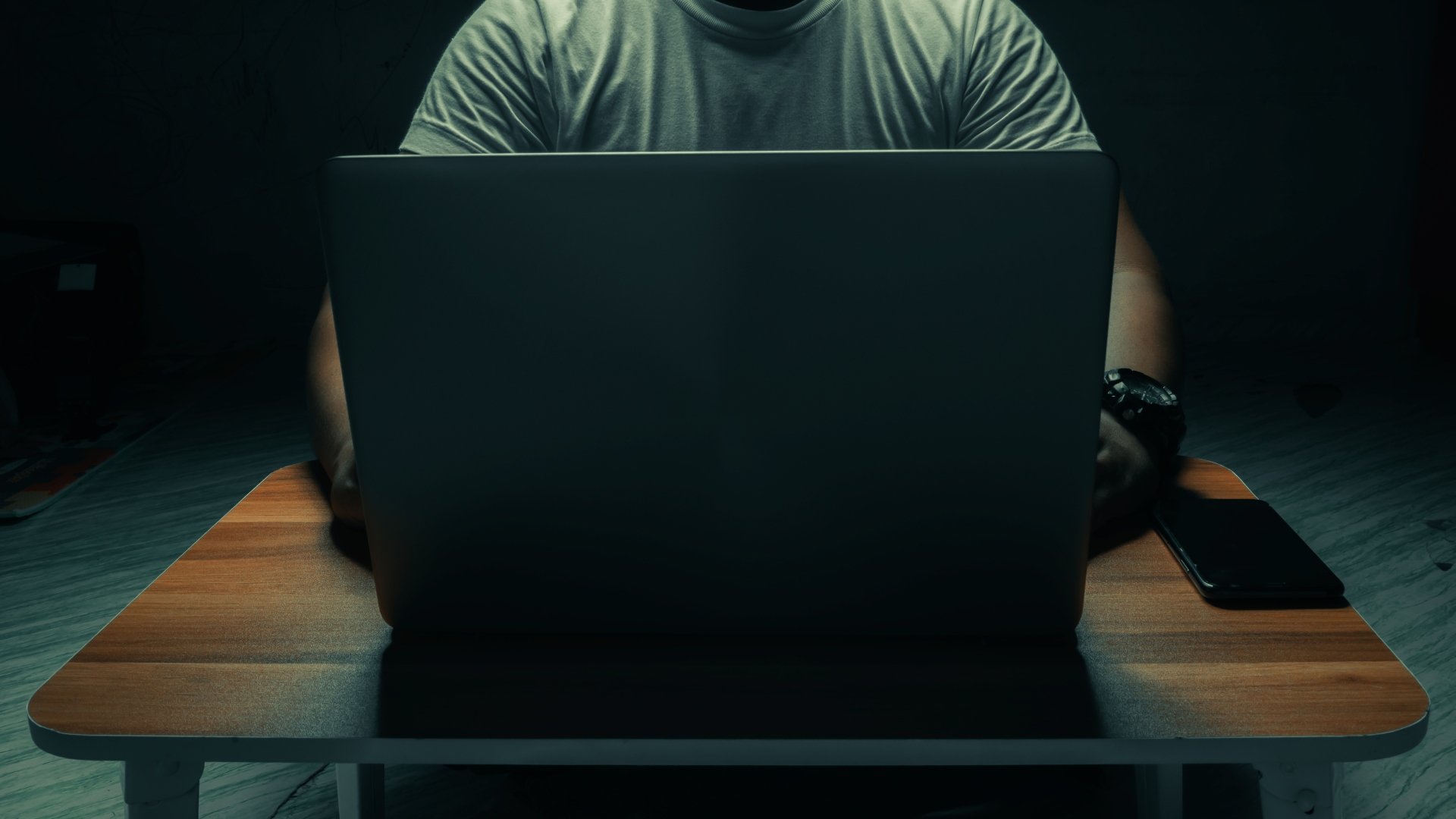 A man in a white shirt sits on a laptop in a dark room. with light