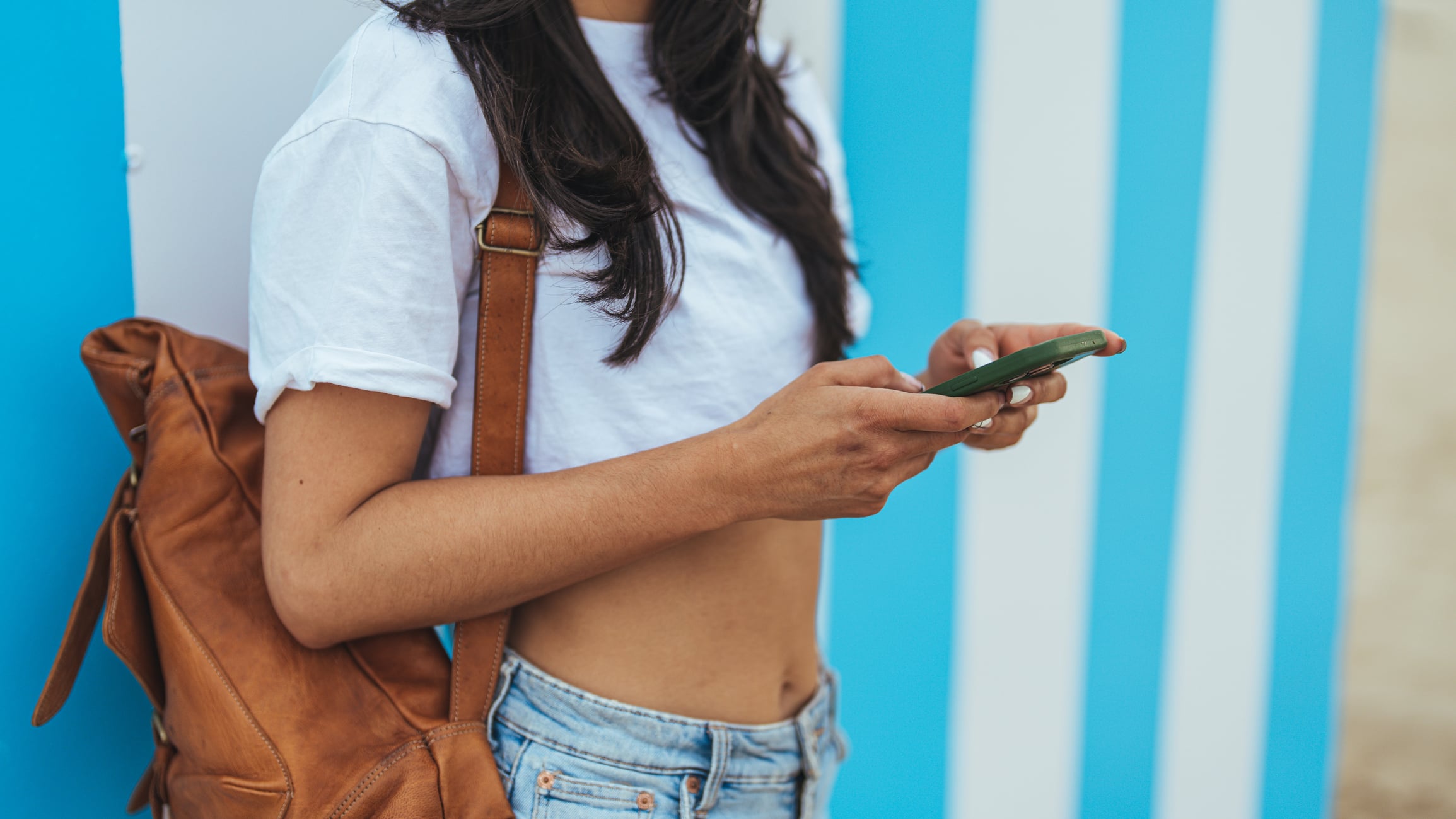 Young woman holds a smartphone while standing outdoors, wearing casual summer clothing and a backpack. The blue striped background adds a vibrant and fresh aesthetic to the scene.