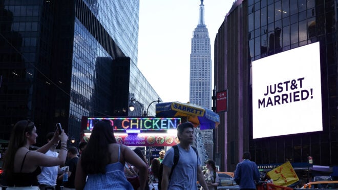 The Empire State Building is seen in the distance as people take photos of a giant screen showing the message "JUST&T MARRIED!"