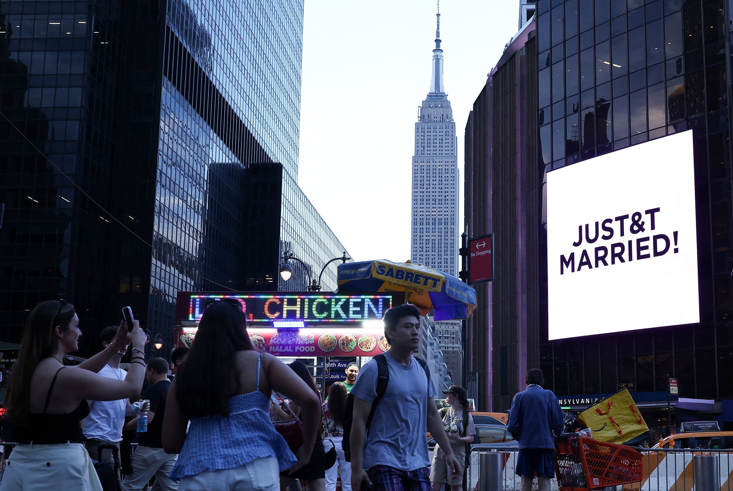 The Empire State Building is seen in the distance as people take photos of a giant screen showing the message "JUST&T MARRIED!"